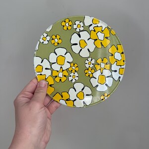 May include: A round, floral-patterned plate featuring a design of white and yellow flowers on a light green background. The plate is held by a person's hand against a neutral gray backdrop. The flowers have a retro, vintage aesthetic.