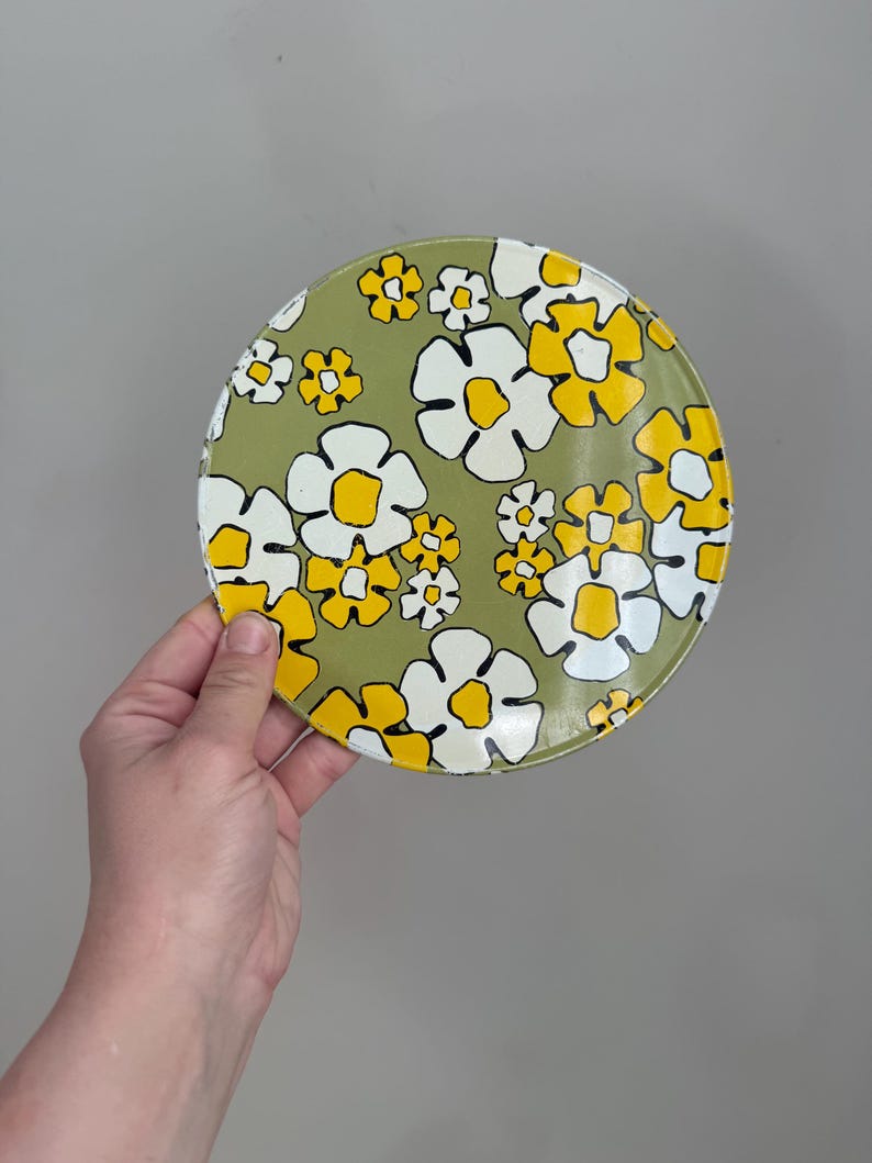 May include: A round, floral-patterned plate featuring a design of white and yellow flowers on a light green background. The plate is held by a person's hand against a neutral gray backdrop. The flowers have a retro, vintage aesthetic.