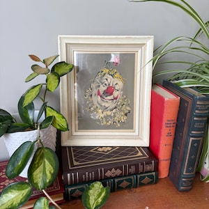 May include: A framed vintage illustration of a clown with a red nose and a pink flower in their hair. The illustration is in a white frame and is sitting on a stack of books.