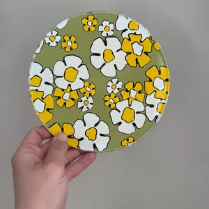 May include: A round, floral-patterned plate with a green background. The plate features white and yellow flowers with black outlines. The plate is held by a hand against a neutral background.
