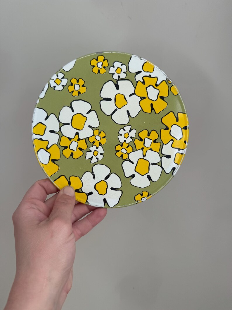 May include: A round, floral-patterned plate with a green background. The plate features white and yellow flowers with black outlines. The plate is held by a hand against a neutral background.