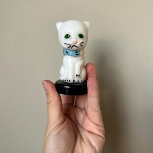 May include: A white cat figurine with green eyes and a blue collar. The cat has black whiskers and is sitting on a black base. The figurine is held in a hand against a neutral background.