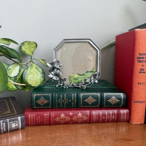Puede incluir: Un marco de fotos octogonal plateado con adornos florales descansa sobre una pila de libros antiguos. Los libros están encuadernados en cuero verde y rojo, con letras doradas y patrones decorativos. Una planta frondosa es visible a la izquierda.