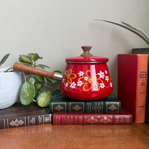 Puede incluir: Una cacerola de esmalte rojo con mango y tapa de madera, decorada con motivos florales blancos y amarillos. La olla vintage se encuentra sobre una pila de libros antiguos con cubiertas verdes y rojas.