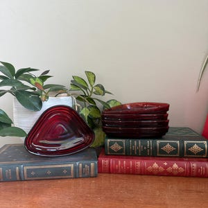 May include: A set of vintage ruby red glass dishes, triangular in shape, stacked on antique books. The books are bound in various colors, including green and red, and are arranged on a wooden surface. A houseplant is visible in the background.