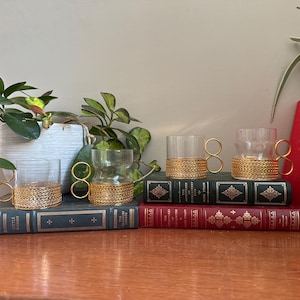 Pode incluir: Quatro xícaras de vidro transparente com alças douradas e faixas douradas tecidas na base. As xícaras estão dispostas sobre livros antigos com capas verdes e vermelhas. As plantas em vasos ao fundo adicionam um toque de vegetação.