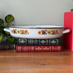 Puede incluir: Una fuente de horno de esmalte blanco con un estampado floral en tonos naranja, amarillo y marrón. La fuente tiene dos asas y un borde marrón. Descansa sobre una pila de libros antiguos con letras doradas.