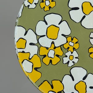 May include: Round metal tray with a floral pattern. The tray features large white flowers with yellow centers and black outlines, set against a green background. The design has a retro, vintage aesthetic.