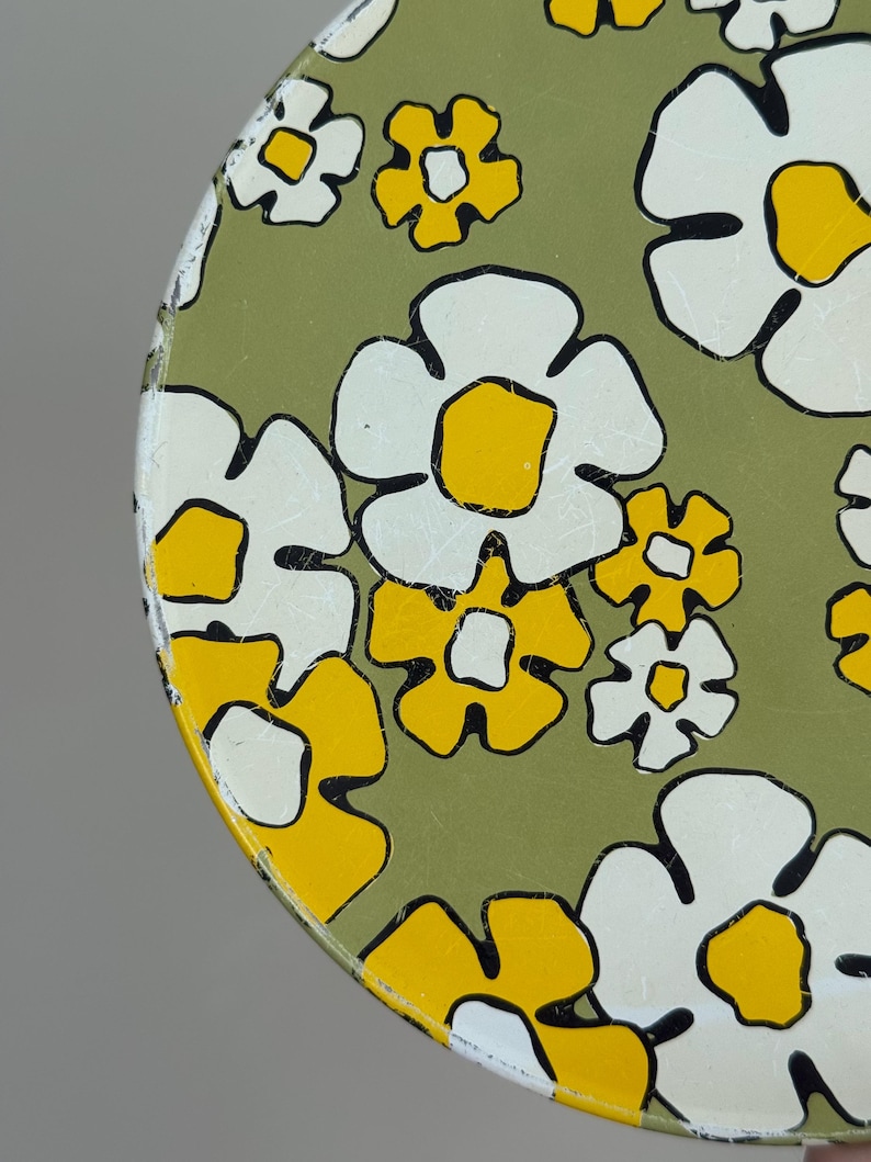 May include: Round metal tray with a floral pattern. The tray features large white flowers with yellow centers and black outlines, set against a green background. The design has a retro, vintage aesthetic.