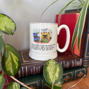 May include: White ceramic mug with a gold rim and a cartoon illustration of a fireplace, a chair, and a cat. The mug has the text "Your supper is on the hearth dear, a pipe and a book that's new, your slippers are by the fire dear, but where the hell are you?"