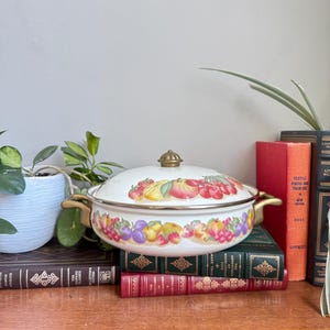 Peut inclure: Casserole en émail blanc avec couvercle, orné d'un motif de fruits colorés. Le plat est doté de poignées dorées et d'un bouton assorti sur le couvercle. Il repose sur une pile de livres anciens, créant une charmante présentation.