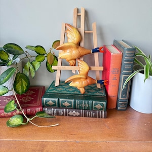 Puede incluir: Dos figuras de patos de cerámica con las alas extendidas, pintadas en tonos amarillos y naranjas, con detalles azules y rojos. Los patos están montados en un pequeño caballete de madera, colocado sobre una pila de libros antiguos.