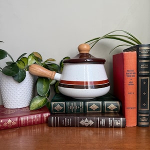 以下が含まれることがあります： 木製の取っ手と蓋が付いた、白と茶色の斑点模様のヴィンテージセラミックソースパン。鍋には茶色とオレンジ色のストライプ模様があります。木製の表面に、アンティークの本や植物と一緒に置かれています。直径約20cm。