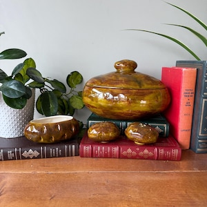 Puede incluir: Un cuenco de cerámica con tapa y tres piezas más pequeñas a juego. Las piezas son marrones con detalles en amarillo y naranja. El cuenco y la tapa tienen forma ovalada. Los artículos se exhiben sobre libros antiguos.