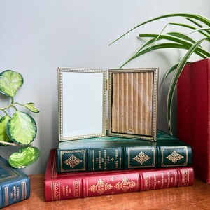 Peut inclure: Un petit cadre photo double, ouvert, de couleur dorée, avec un intérieur blanc et un intérieur marron, posé sur une pile de livres anciens. Les livres sont verts et rouges avec des lettres et des motifs dorés. Une plante est en arrière-plan.