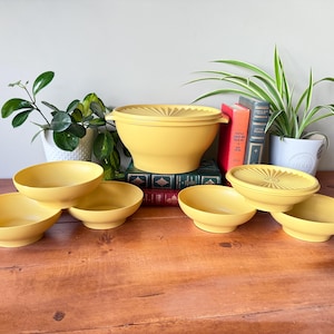 Op de afbeelding: Een verzameling vintage gele plastic kommen en containers. De set bevat kommen in verschillende maten, sommige met deksels, en een grotere container met een deksel. De items zijn gerangschikt op een houten oppervlak, met planten en boeken op de achtergrond.