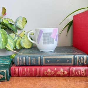 Peut inclure: Tasse en céramique blanche avec un motif géométrique lavande et rose, posée sur une pile de livres anciens. Les livres sont reliés en cuir vert et rouge avec des lettres dorées. Des plantes vertes sont en arrière-plan.