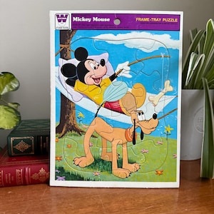 Puede incluir: Rompecabezas vintage de Mickey Mouse con Mickey Mouse en una hamaca y Pluto. El rompecabezas tiene una ilustración colorida con un fondo de árbol y hierba. La caja dice "Mickey Mouse" y "FRAME-TRAY PUZZLE".
