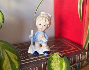 Op de afbeelding: Een porseleinen beeldje van een meisje dat een blauwe jurk draagt en een bezem vasthoudt. Het beeldje staat op een stapel boeken met bruine en groene omslagen.