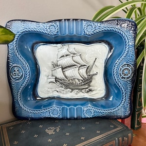 May include: A rectangular blue ceramic ashtray with a raised relief of a sailing ship. The ashtray has a rope design around the edges and features an anchor and a ship's wheel. The center is white with a detailed ship.