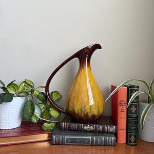 Op de afbeelding: Een vintage keramische kan met een lange handgreep, met een kleurovergang van bruin naar geel. De kan staat op een stapel boeken, met een plant in een witte pot links en andere boeken rechts.