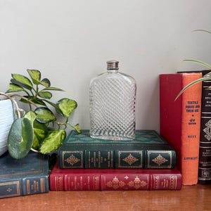 Può includere: Una fiaschetta di vetro trasparente e rettangolare con tappo argentato, caratterizzata da un motivo a losanghe. Poggia su una pila di libri antichi con copertine verdi e rosse, creando un'estetica vintage. Una piccola pianta è visibile a sinistra.