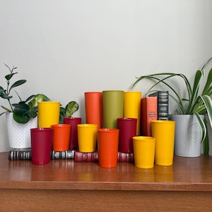 Puede incluir: Una colección de vasos cilíndricos de colores en tonos amarillo, naranja, rojo y verde. Los vasos están colocados sobre una superficie de madera con libros y plantas en macetas en el fondo. Los vasos varían en altura y diámetro.