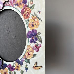 Puede incluir: Un marco de fotos de madera blanco con un dise&ntilde;o floral con pensamientos morados, amarillos y naranjas. Una peque&ntilde;a mariposa amarilla se encuentra en la esquina inferior derecha del marco.