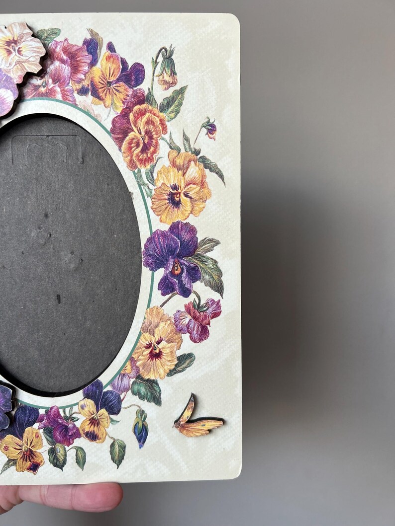 Puede incluir: Un marco de fotos de madera blanco con un dise&ntilde;o floral con pensamientos morados, amarillos y naranjas. Una peque&ntilde;a mariposa amarilla se encuentra en la esquina inferior derecha del marco.