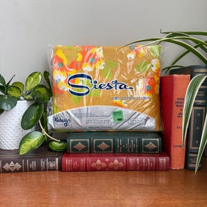 Könnte beinhalten: Ein verpacktes Siesta Twin-Top-Flachbettlaken mit einem Blumenmuster in Orange, Gelb und Weiß. Das Laken ist mit 100 % Baumwolle gekennzeichnet. Das Paket liegt auf einem Stapel alter Bücher.