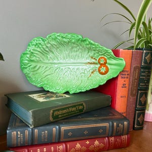 Puede incluir: Un plato verde de cerámica con forma de lechuga con dos acentos de tomate naranja. El plato descansa sobre una pila de libros antiguos con letras doradas y cubiertas rojas, verdes y naranjas.
