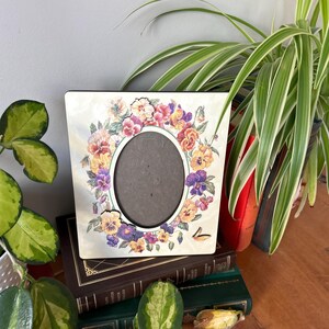 Puede incluir: Un marco de fotos blanco con un dise&ntilde;o floral con pensamientos morados, amarillos y naranjas. El marco tiene una abertura ovalada para una foto.
