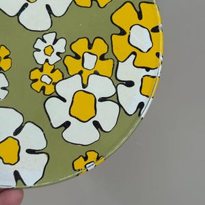 May include: A round, vintage metal container with a floral pattern. The design features large white flowers with yellow centers and black outlines, set against a muted green background. The container's edge is visible.