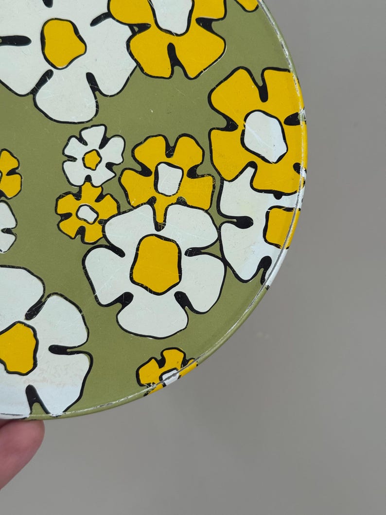 May include: A round, vintage metal container with a floral pattern. The design features large white flowers with yellow centers and black outlines, set against a muted green background. The container's edge is visible.