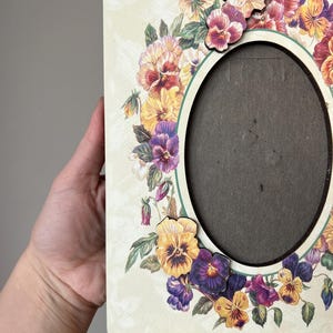 Puede incluir: Un marco de fotos de madera blanco con una abertura ovalada. El marco est&aacute; decorado con un borde de pensamientos coloridos.