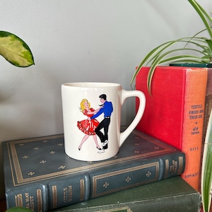 Peut inclure: Une tasse en céramique blanche avec une illustration colorée d'un couple dansant. La femme porte une robe rouge et l'homme porte une chemise bleue et un pantalon noir.