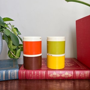 Puede incluir: Dos recipientes cilíndricos de plástico con tapa. El recipiente de la izquierda presenta una sección superior de color naranja brillante y una inferior de color marrón oscuro. El de la derecha tiene una sección superior verde claro y una base amarilla.