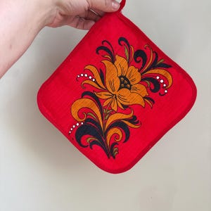 May include: A square red potholder featuring a central floral design in shades of orange, black, and white. The fabric has a subtle textured weave, and a small red fabric loop is attached to the top corner for hanging purposes.