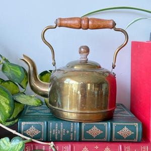 Peut inclure: Théière vintage en laiton avec une poignée et un bouton en bois. La théière a un bec courbé et repose sur une pile de livres avec des lettres dorées et des motifs décoratifs. Les livres sont verts et rouges.
