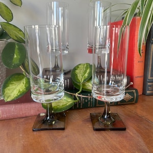 May include: Set of four clear glass drinking glasses with a square base and a green stem. The glasses are arranged on a wooden surface.