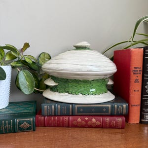 May include: A decorative ceramic jar with a lid, featuring a white and green color scheme. The jar is placed on a stack of vintage books with red, green, and gold covers. The books are arranged on a wooden surface.