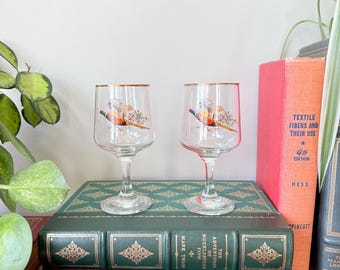 May include: Two clear wine glasses with gold rims and a colorful floral design. The glasses are displayed on a stack of vintage books with green and brown covers. A red book is visible in the background.