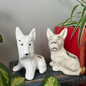 May include: Two white ceramic dog planter figurines. The dogs are sitting and have black painted eyes and noses. The planters have a gold rim.