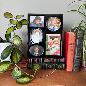 May include: A black picture frame with six openings, holding photos of different people. The frame is sitting on a stack of books.