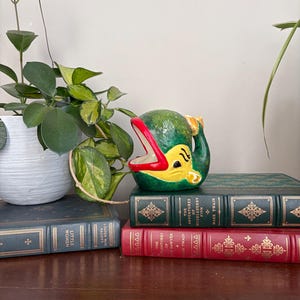 Può includere: Una statuetta di balena in ceramica con corpo verde, ventre giallo e una bocca spalancata con labbra rosso brillante. La balena presenta una piccola pinna caudale e dettagli oculari neri. È posizionata sopra due libri con copertina rigida.
