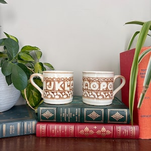 May include: Two off-white ceramic mugs featuring a brown embossed alphabet pattern. The left mug displays the letters I, J, K, L, and M, while the right mug shows N, O, P, Q, and R. Each mug has a curved handle and a cylindrical shape.