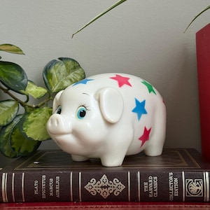 Może przedstawiać: Biała ceramiczna skarbonka z kolorowymi gwiazdami. Świnka stoi na stosie książek ze złotymi literami. Książki są w różnych kolorach, w tym czerwonym i brązowym.