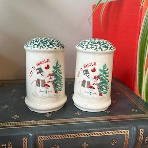 May include: A pair of white ceramic salt and pepper shakers with green, red, and black details. Each shaker has a snowman family design and the text "Let it Snow!". The tops of the shakers have a green, textured pattern.