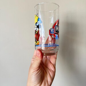 May include: A clear, vintage drinking glass featuring a cartoon character design. The glass has a blue stripe near the base and a colorful graphic of a character with the letters "YM" in a red and blue emblem. The glass is held up.