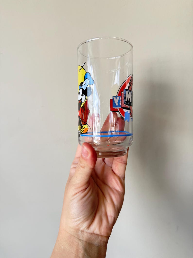 May include: A clear, vintage drinking glass featuring a cartoon character design. The glass has a blue stripe near the base and a colorful graphic of a character with the letters "YM" in a red and blue emblem. The glass is held up.
